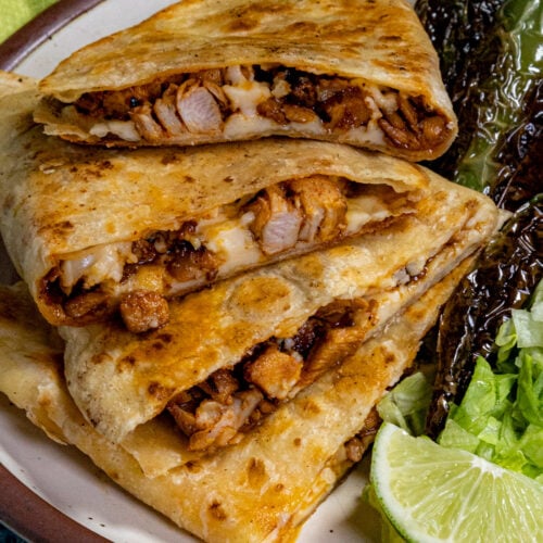 A plate with three stacked homemade Chicken Quesadillas, a side of grilled peppers, shredded lettuce, and lime wedges on a brown-rimmed dish against a blue background.