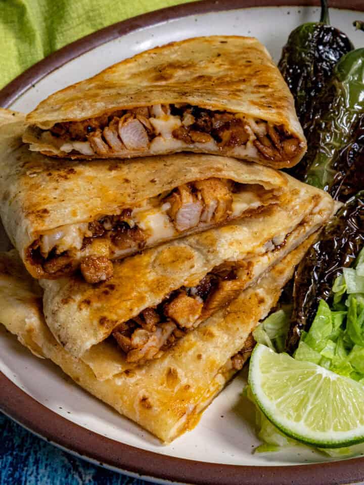 A plate with three stacked homemade Chicken Quesadillas, a side of grilled peppers, shredded lettuce, and lime wedges on a brown-rimmed dish against a blue background.
