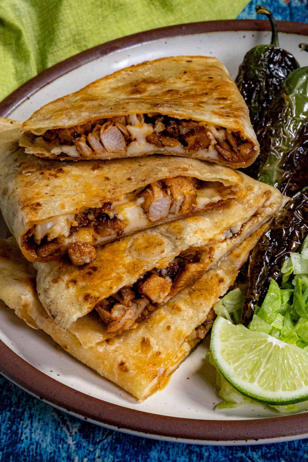 A plate with three stacked homemade Chicken Quesadillas, a side of grilled peppers, shredded lettuce, and lime wedges on a brown-rimmed dish against a blue background.