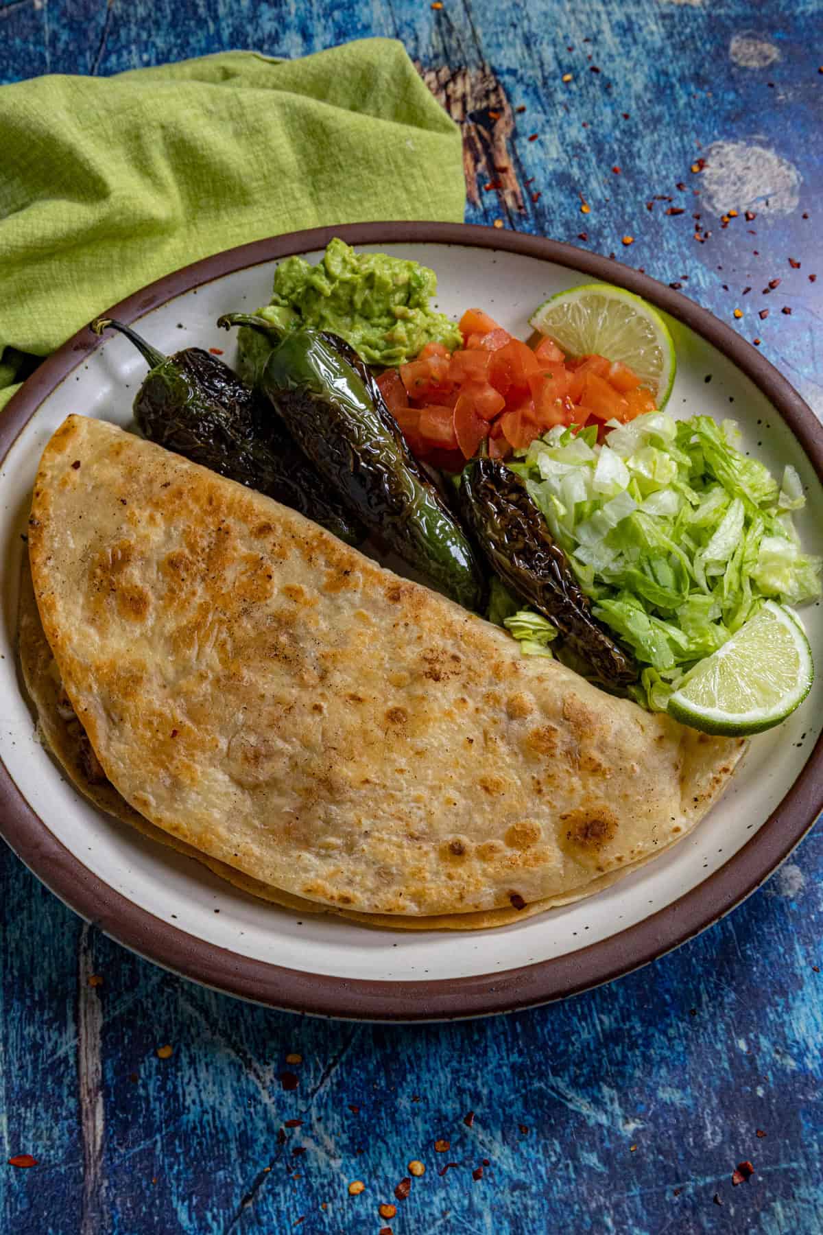 A plate with a large folded restaurant-style quesadilla, grilled jalape&ntilde;os, guacamole, chopped tomatoes, shredded lettuce, and lime wedges sits on a blue rustic table with a green napkin.