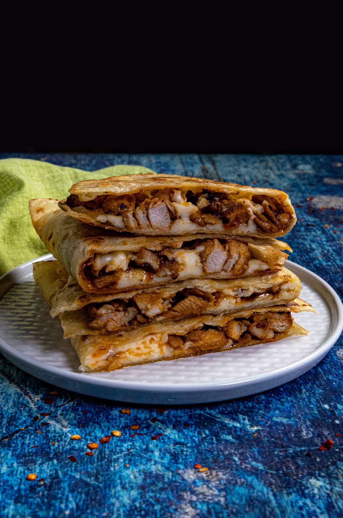 A stack of four folded Chicken and Cheese Quesadillas filled with grilled chicken and melted cheese sits on a white plate. The plate rests on a blue textured surface with a green cloth in the background.