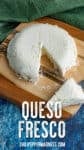 A round wheel of crumbly white cheese, queso fresco, with a wedge cut out, sits on a wooden cutting board. A knife holds the wedge in front of the cheese. A green cloth is in the background, and a blue surface is underneath.
