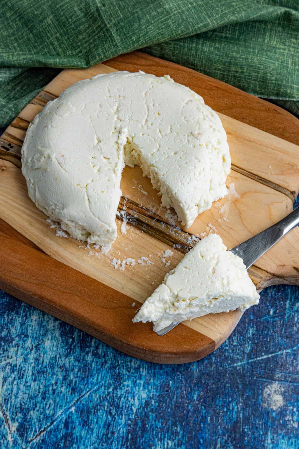A round wheel of crumbly white cheese, queso fresco, with a wedge cut out, sits on a wooden cutting board. A knife holds the wedge in front of the cheese. A green cloth is in the background, and a blue surface is underneath.