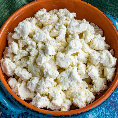 A clay bowl filled with crumbled fresh homemade queso fresco sits on a blue textured surface, with a green cloth partially visible in the background.
