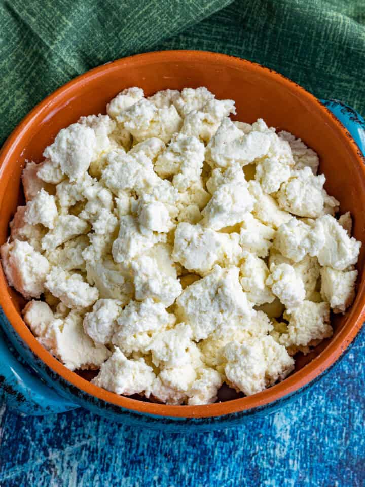 A clay bowl filled with crumbled fresh homemade queso fresco sits on a blue textured surface, with a green cloth partially visible in the background.