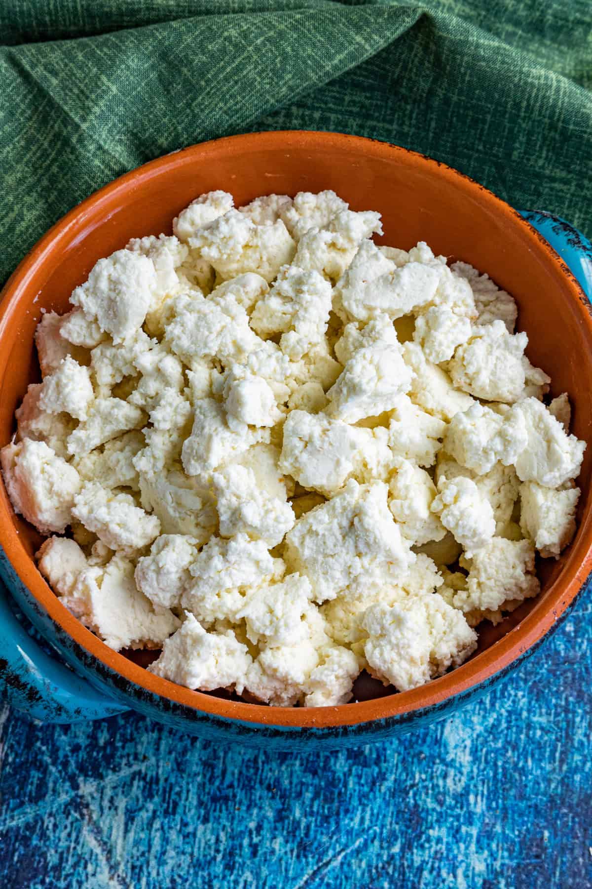 A clay bowl filled with crumbled fresh homemade queso fresco sits on a blue textured surface, with a green cloth partially visible in the background.