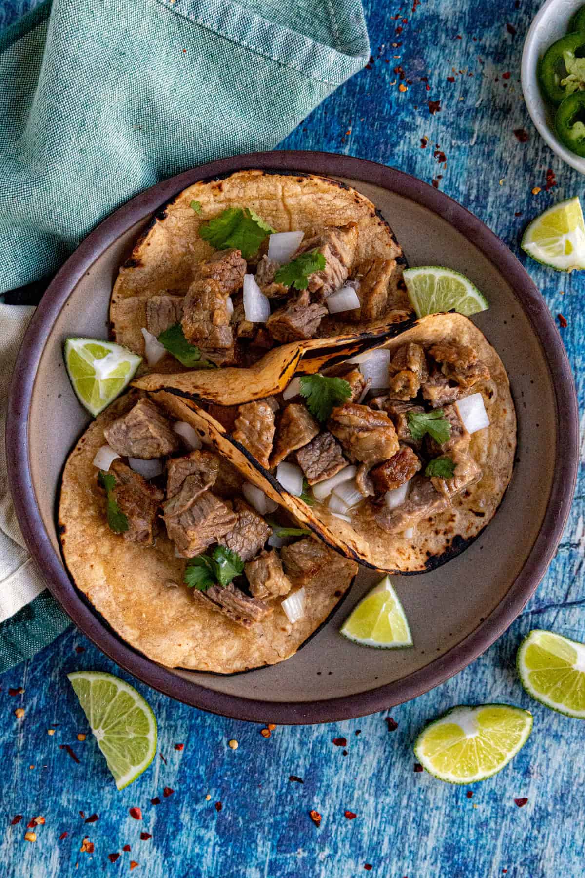 Three Suadero Tacos on a plate with chopped onions and cilantro, served on corn tortillas. Lime wedges are placed on the plate and around it. The background is a rustic blue surface with a green cloth napkin nearby.