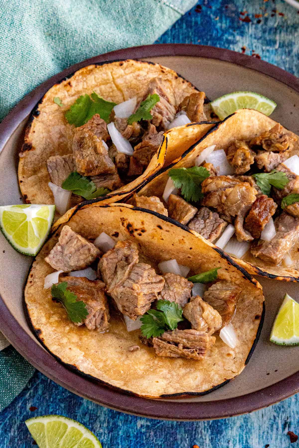 Three corn tortillas filled with chunks of pork, chopped onions, and cilantro are arranged on a plate as flavorful Suadero Tacos, garnished with lime wedges on a blue textured surface.