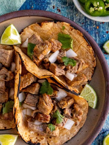 Three corn tortillas filled with chopped beef, diced onions, and cilantro sit on a plate with lime wedges. Nearby, a bowl of sliced jalapeños and extra lime wedges complement these Suadero Tacos on a blue textured surface.