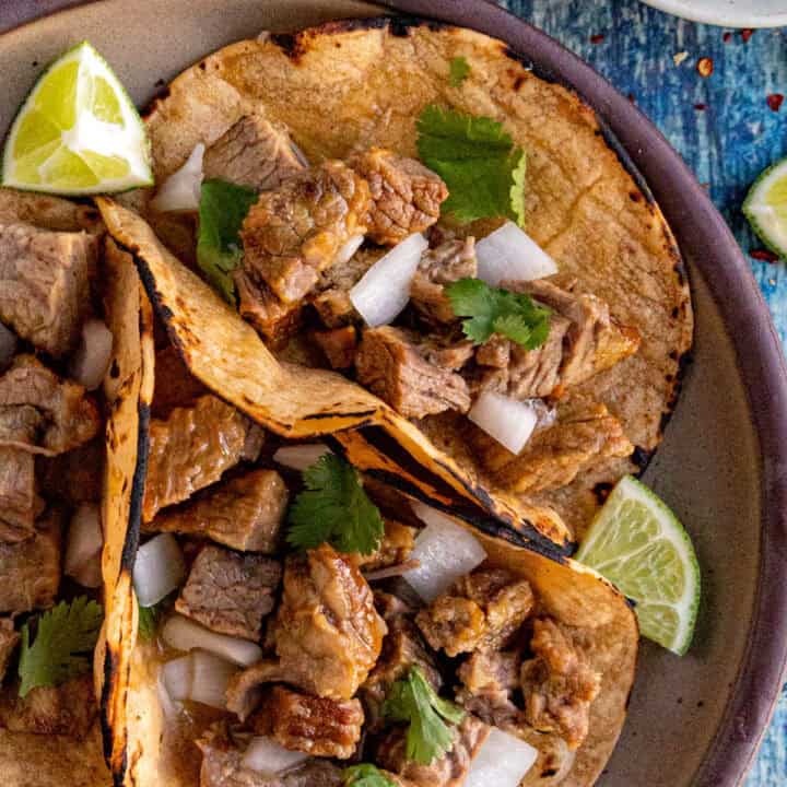 Three corn tortillas filled with chopped beef, diced onions, and cilantro sit on a plate with lime wedges. Nearby, a bowl of sliced jalapeños and extra lime wedges complement these Suadero Tacos on a blue textured surface.
