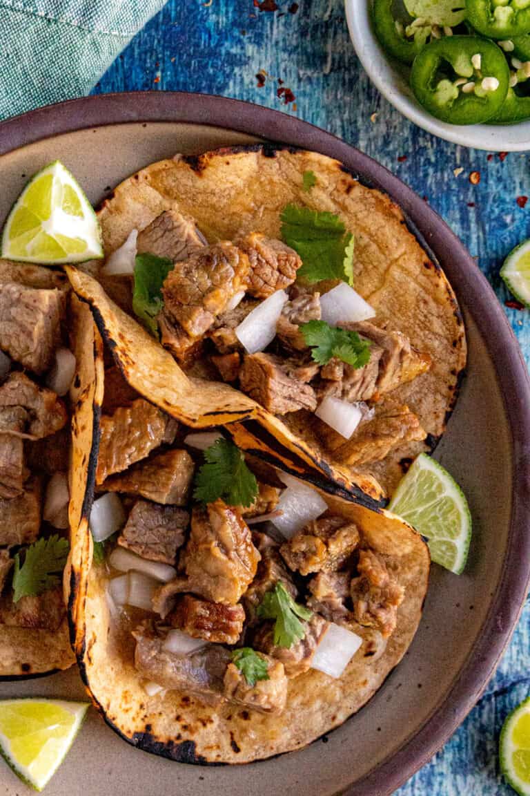 Three corn tortillas filled with chopped beef, diced onions, and cilantro sit on a plate with lime wedges. Nearby, a bowl of sliced jalape&ntilde;os and extra lime wedges complement these Suadero Tacos on a blue textured surface.