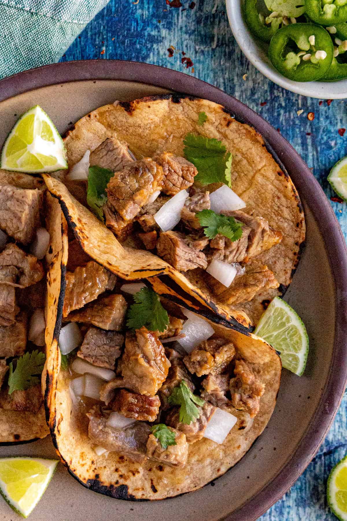 Three corn tortillas filled with chopped beef, diced onions, and cilantro sit on a plate with lime wedges. Nearby, a bowl of sliced jalapeños and extra lime wedges complement these Suadero Tacos on a blue textured surface.