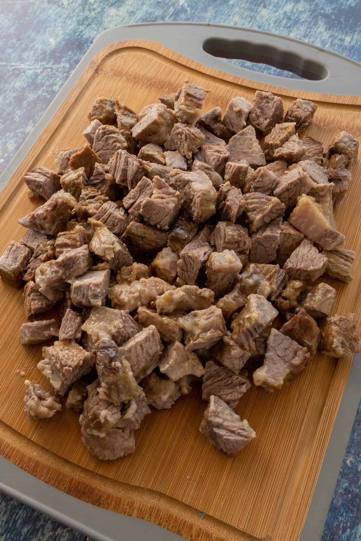 A pile of cooked beef cubes, perfect for Suadero Tacos, sits on a wooden cutting board with a gray handle, set on a blue marbled surface.