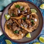 Three Suadero Tacos topped with chopped onions and cilantro are served on toasted corn tortillas with lime wedges. The plate rests on a blue textured surface, with extra lime wedges nearby for a fresh finish.