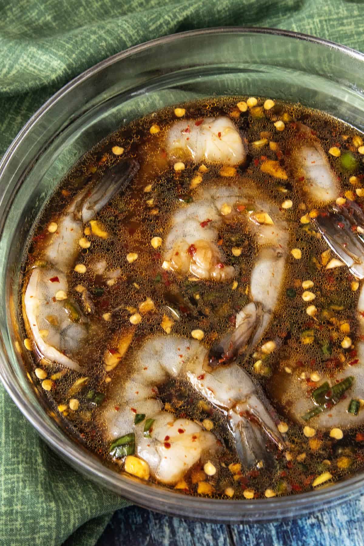 Raw shrimp marinating in a glass bowl with a dark, homemade Teriyaki Marinade, chopped garlic, red pepper flakes, and herbs, set on a green cloth background.