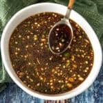 A bowl of dark teriyaki marinade sauce with visible chopped garlic, green onions, and red pepper flakes, with a spoon resting inside. The bowl sits on a green cloth and a blue wooden surface.