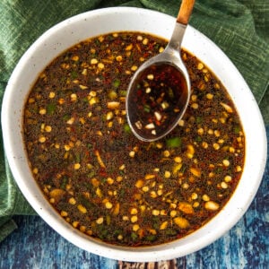 A bowl of dark teriyaki marinade sauce with visible chopped garlic, green onions, and red pepper flakes, with a spoon resting inside. The bowl sits on a green cloth and a blue wooden surface.