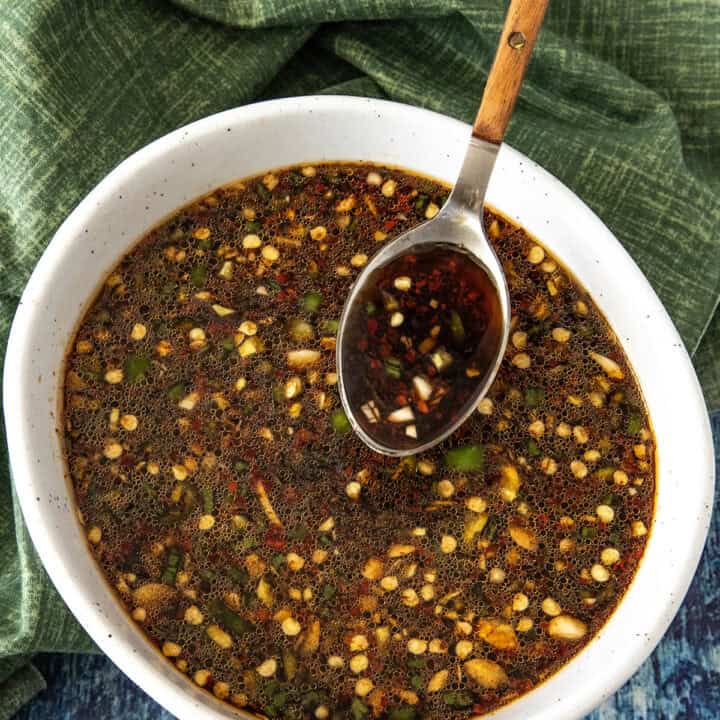 A white bowl filled with a dark, chunky homemade Teriyaki Marinade dipping sauce, featuring chopped garlic, chili, and herbs. A wooden-handled spoon rests in the bowl, with a green cloth and blue wooden surface underneath.