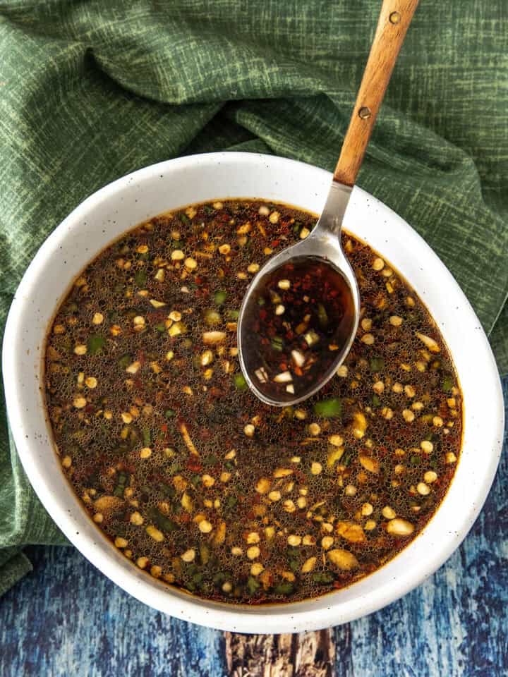 A white bowl filled with a dark, chunky homemade Teriyaki Marinade dipping sauce, featuring chopped garlic, chili, and herbs. A wooden-handled spoon rests in the bowl, with a green cloth and blue wooden surface underneath.