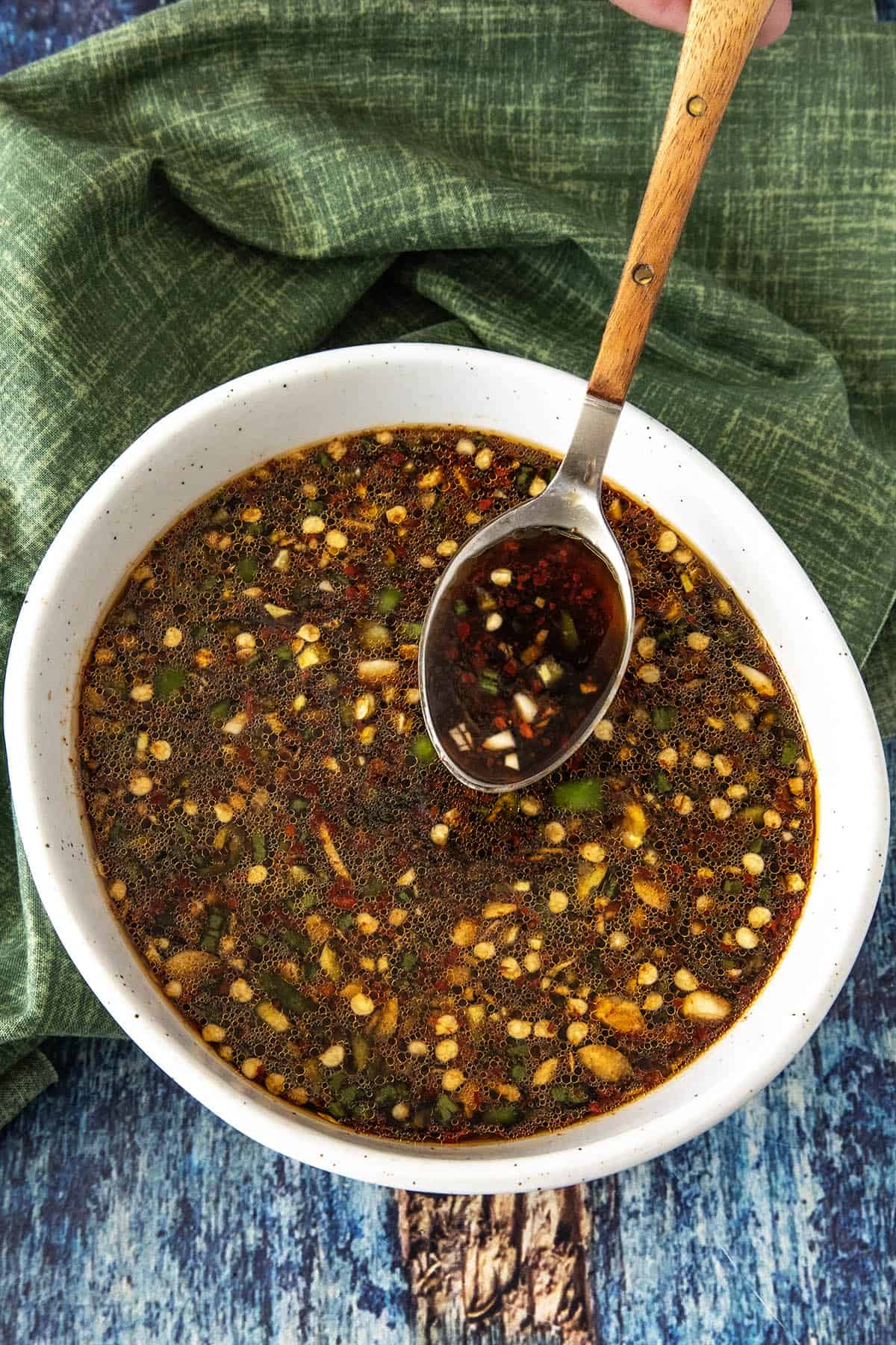 A white bowl filled with a dark, chunky homemade Teriyaki Marinade dipping sauce, featuring chopped garlic, chili, and herbs. A wooden-handled spoon rests in the bowl, with a green cloth and blue wooden surface underneath.