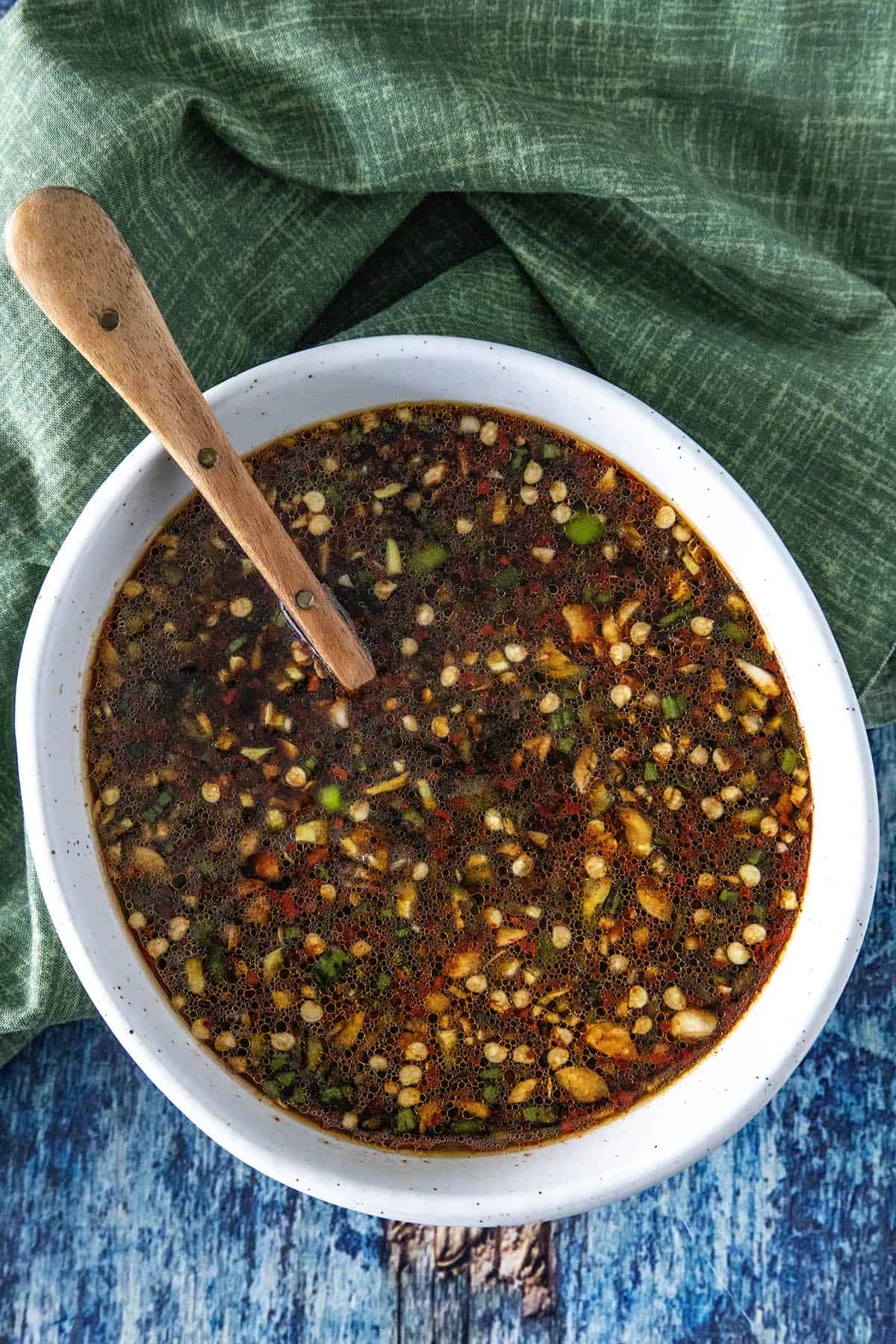 A white bowl filled with a dark, chunky easy Teriyaki Marinade featuring chopped herbs, seeds, and spices. A wooden spoon rests in the bowl, with a green cloth and blue wooden surface in the background.