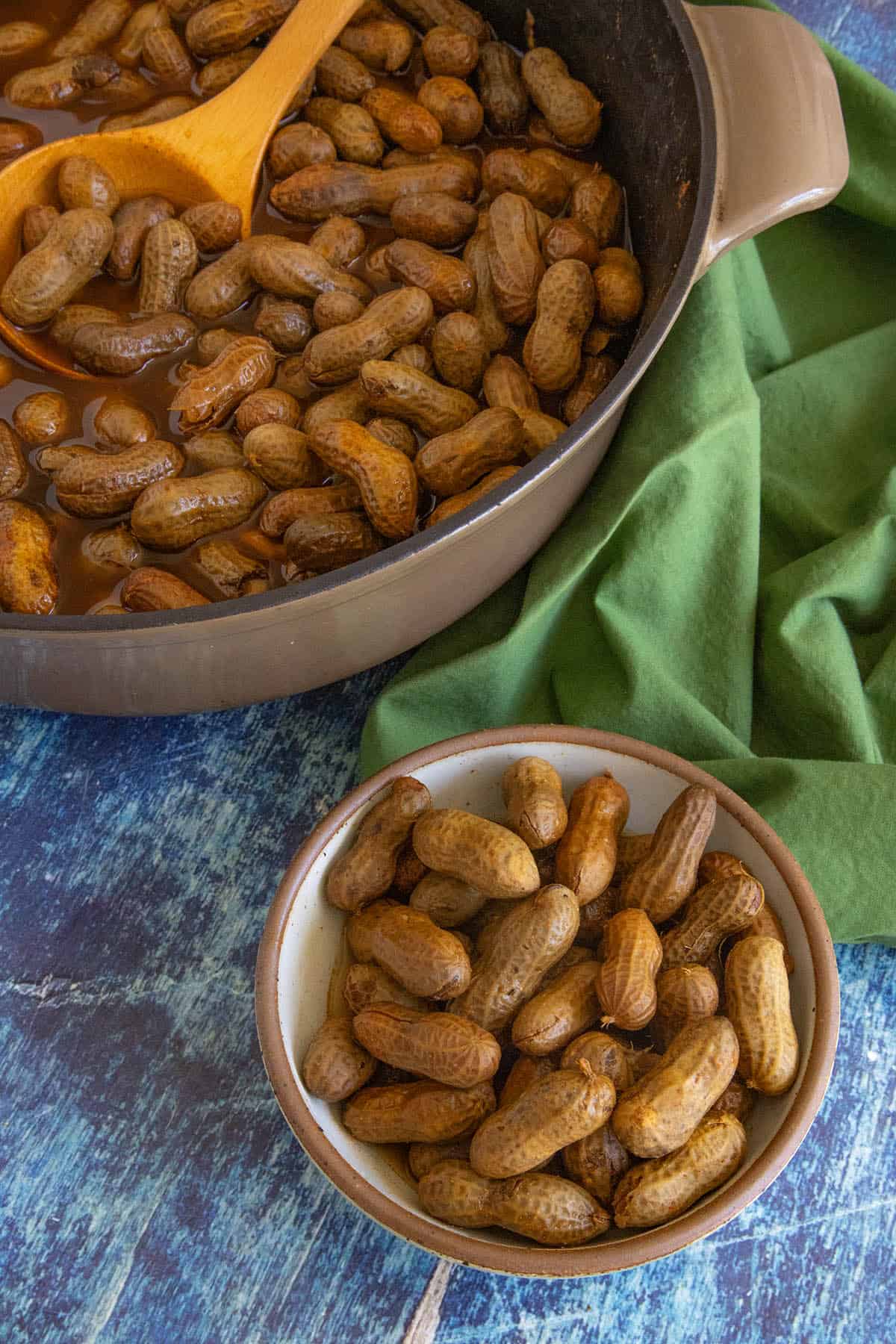 A bowl of Cajun Boiled Peanuts sits on a blue wooden surface next to a large pot filled with more spicy boiled peanuts and a wooden spoon, with a green cloth nearby.
