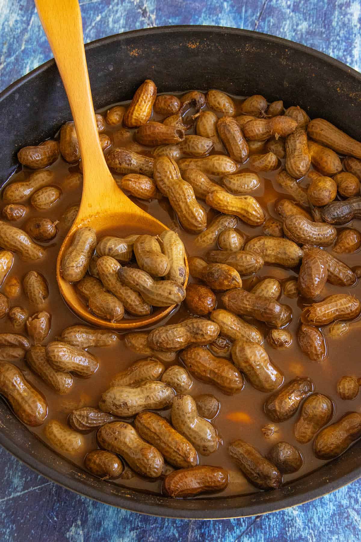 A pot filled with Cajun Boiled Peanuts in a brown, seasoned liquid. A wooden spoon scoops up some of the flavorful peanuts, and the pot rests on a blue textured surface.
