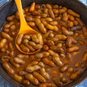 A pot filled with spicy Cajun Boiled Peanuts in their shells, sitting in a brown, seasoned liquid. A wooden spoon is scooping some peanuts from the pot.