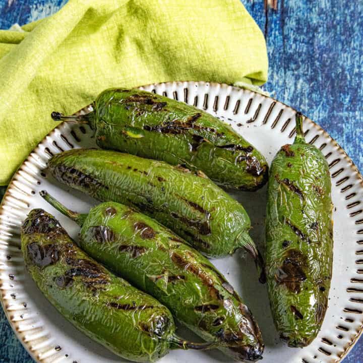 Chiles toreados recipe with five charred blistered jalapenos on a plate, drizzled with lime, on a blue wooden surface.