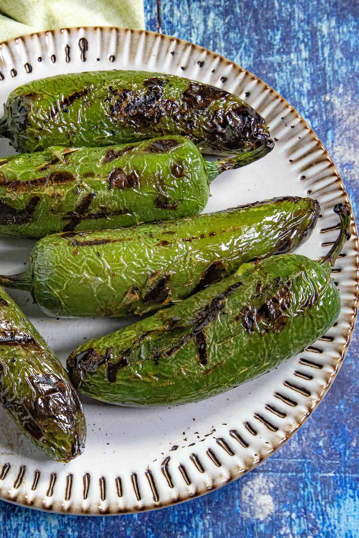 A plate with several charred, grilled green Chiles Toreados sits on a blue surface. The peppers have blistered, blackened skin from roasting.