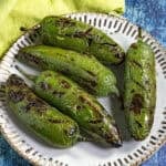 A plate of six Chiles Toreados, their shiny green skins charred with black marks, rests on a white dish with a decorative rim, set against a blue background and a yellow cloth.