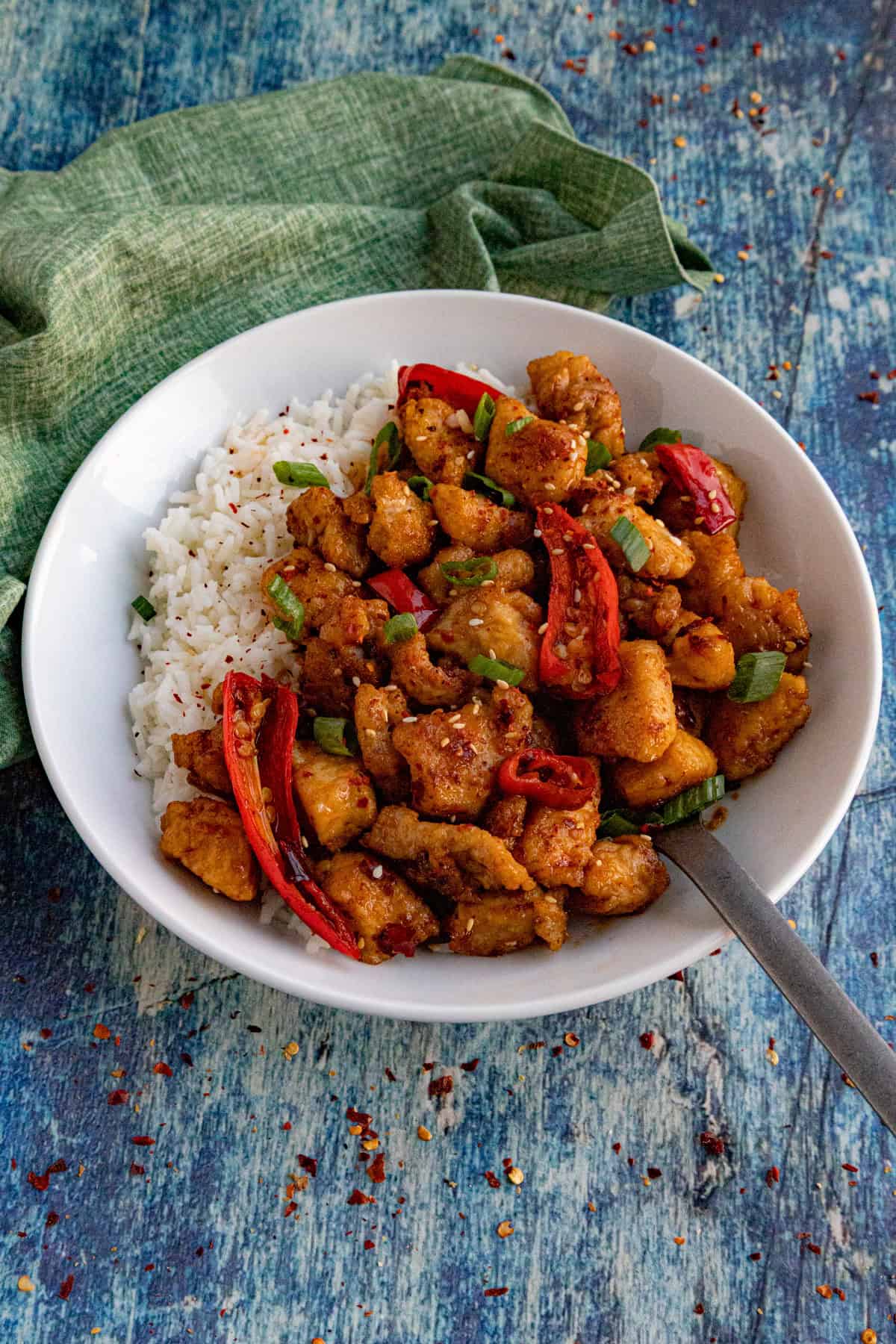 A white bowl filled with white rice and spicy Chili Crisp Chicken, garnished with sliced red peppers and green onions, sits on a rustic blue wooden surface with a green cloth nearby.