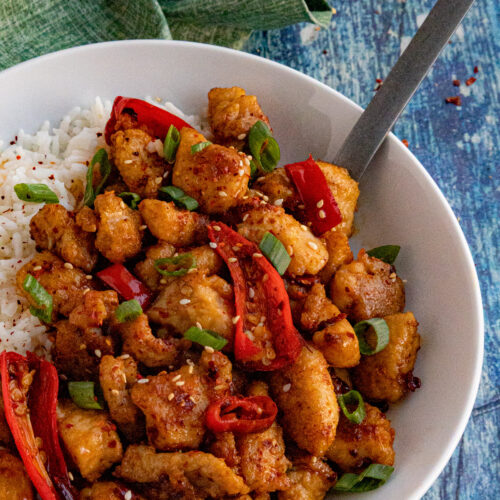 A white bowl filled with white rice and Chili Crisp Chicken, topped with sliced red chilies and green onions, with a fork resting inside; all on a blue textured surface with a green cloth nearby.