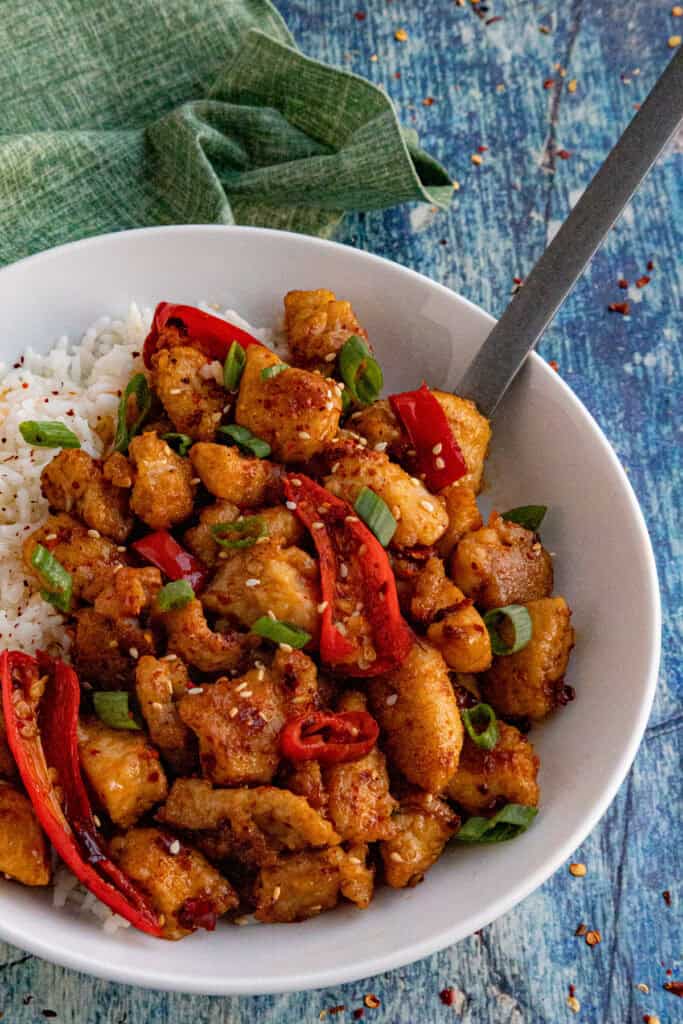 A white bowl filled with white rice and Chili Crisp Chicken, topped with sliced red chilies and green onions, with a fork resting inside; all on a blue textured surface with a green cloth nearby.