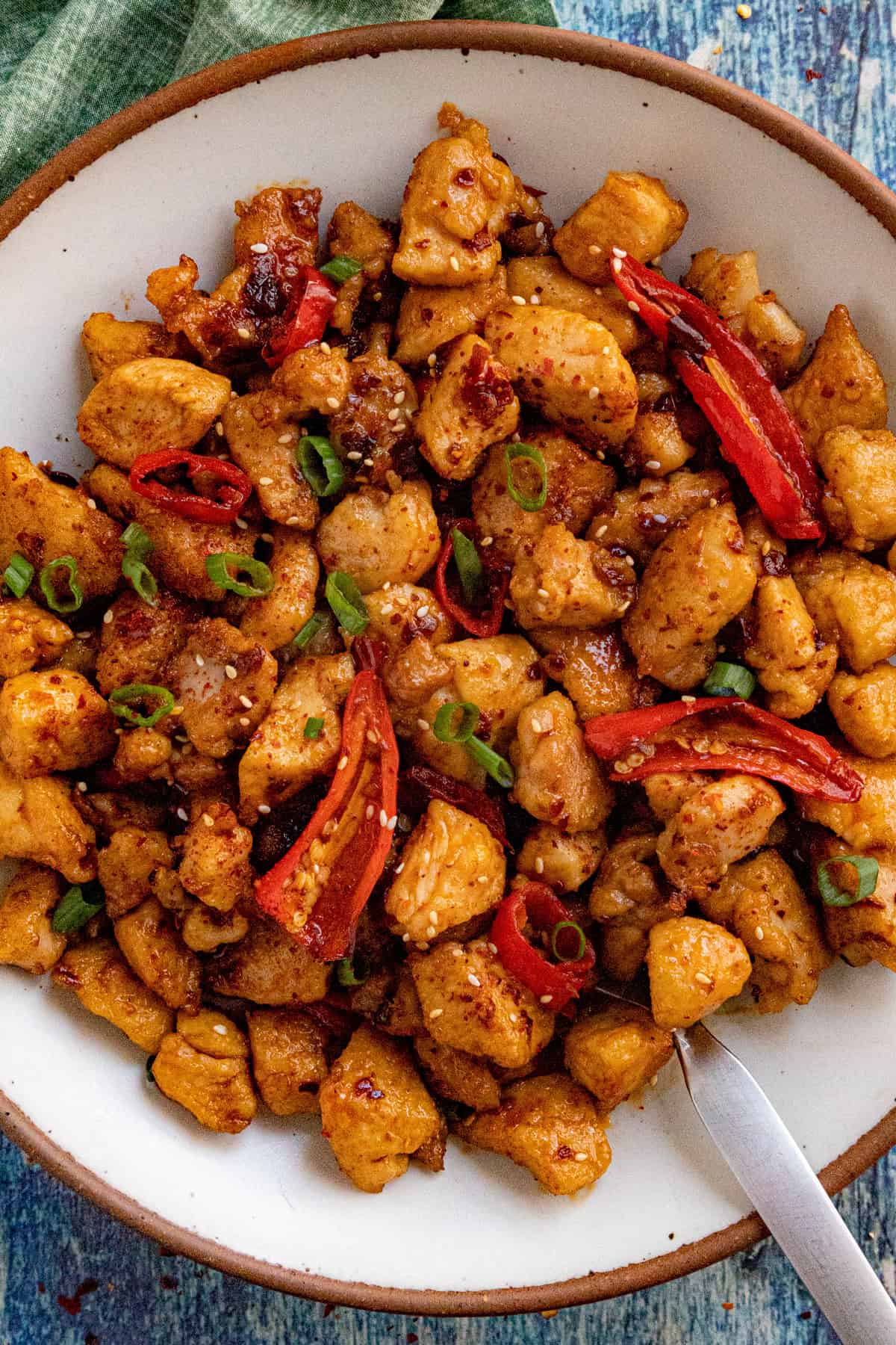 A bowl of spicy chili crisp chicken garnished with sliced red chili peppers, chopped green onions, and sesame seeds, served with a spoon on a blue textured surface.