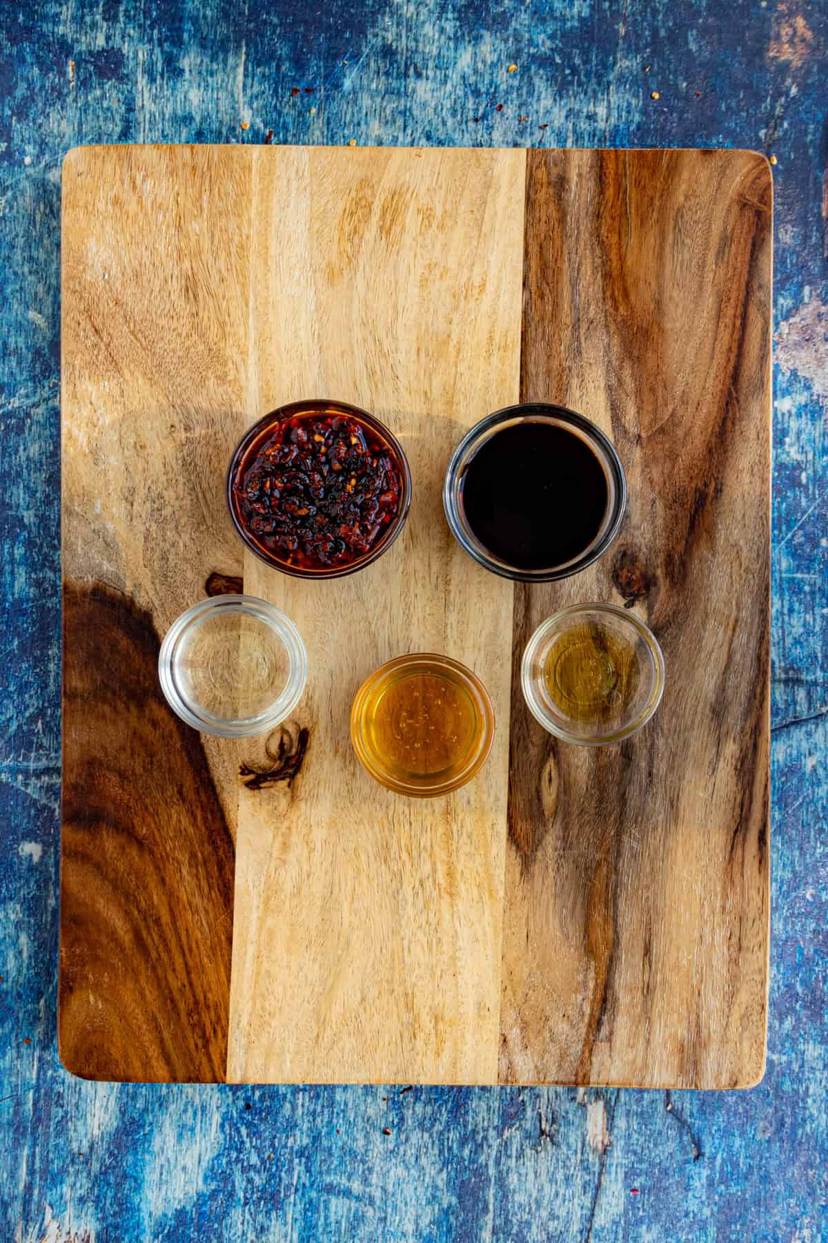 Five small glass bowls containing various chili crisp chicken ingredients&mdash;like chili paste, soy sauce, and oils&mdash;are arranged on a wooden cutting board placed on a blue textured surface.