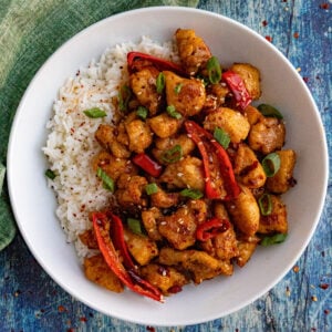 A white bowl filled with white rice and easy chili crisp chicken, garnished with sliced red chilies, green onions, and sesame seeds, on a blue textured surface.