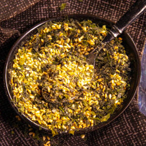 A black bowl filled with homemade furikake seasoning&mdash;a blend of seeds, dried seaweed flakes, and spices&mdash;rests on a dark patterned cloth, with a small spoon partially submerged in the savory mix.