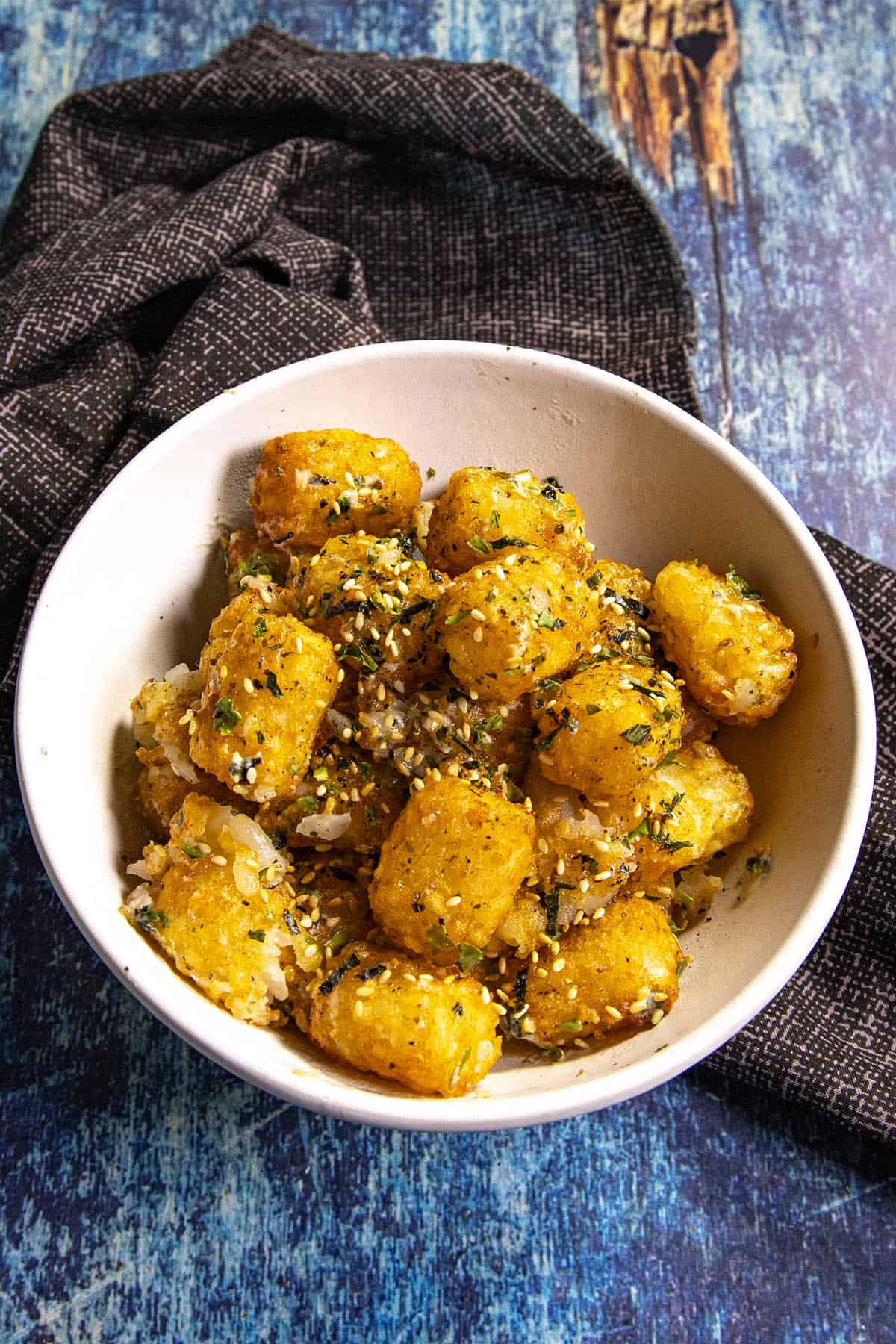 A white bowl filled with golden-brown tater tots sprinkled with herbs and seasonings, inspired by the crunch of furikake fries, sits on a blue wooden surface next to a dark textured cloth.