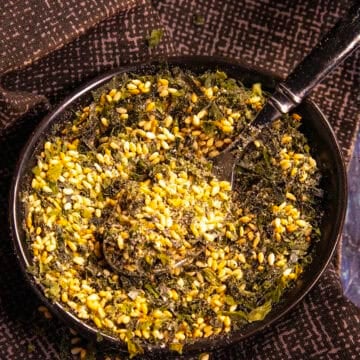 A black bowl filled with homemade furikake seasoning—a blend of seeds, herbs, and spices—rests on a dark patterned cloth, with a black spoon inside.