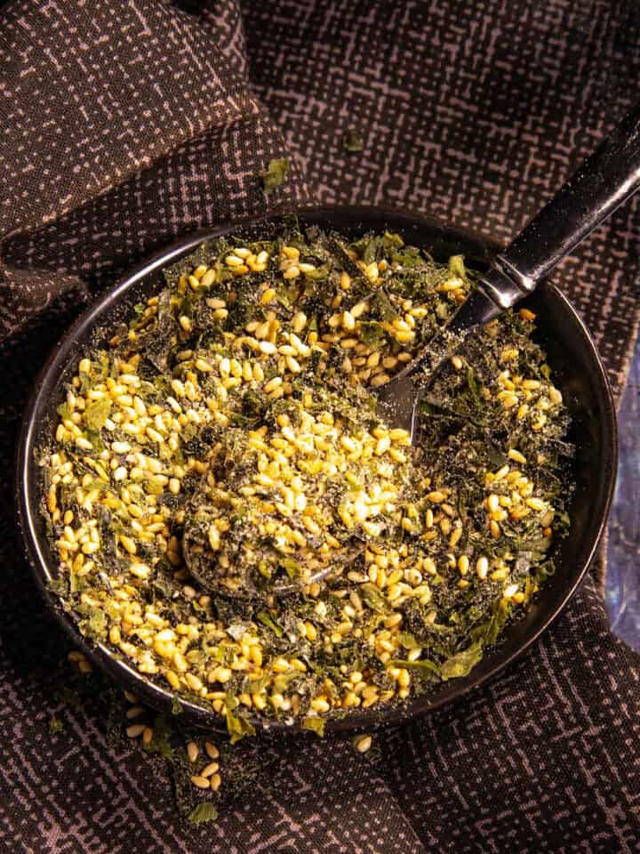 A black bowl filled with homemade furikake seasoning—a blend of seeds, herbs, and spices—rests on a dark patterned cloth, with a black spoon inside.