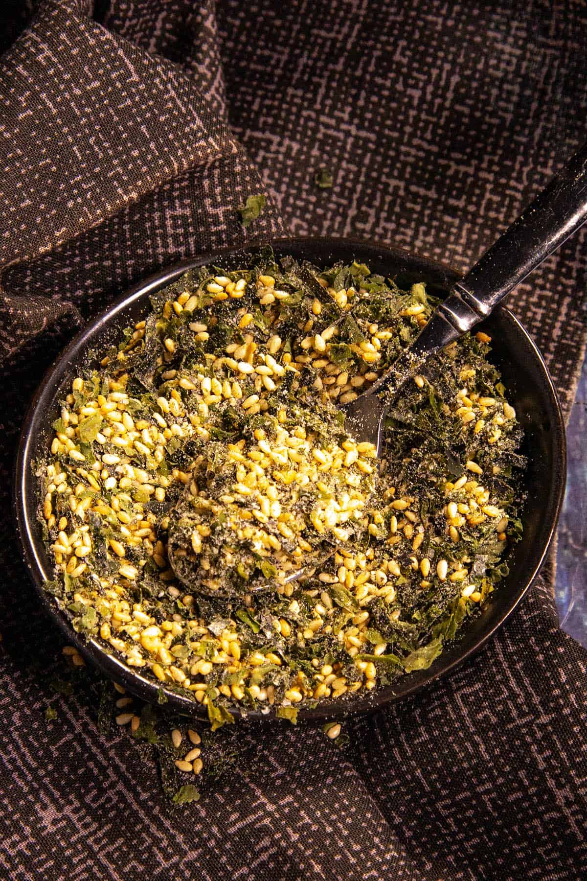 A black bowl filled with homemade furikake seasoning—a blend of seeds, herbs, and spices—rests on a dark patterned cloth, with a black spoon inside.