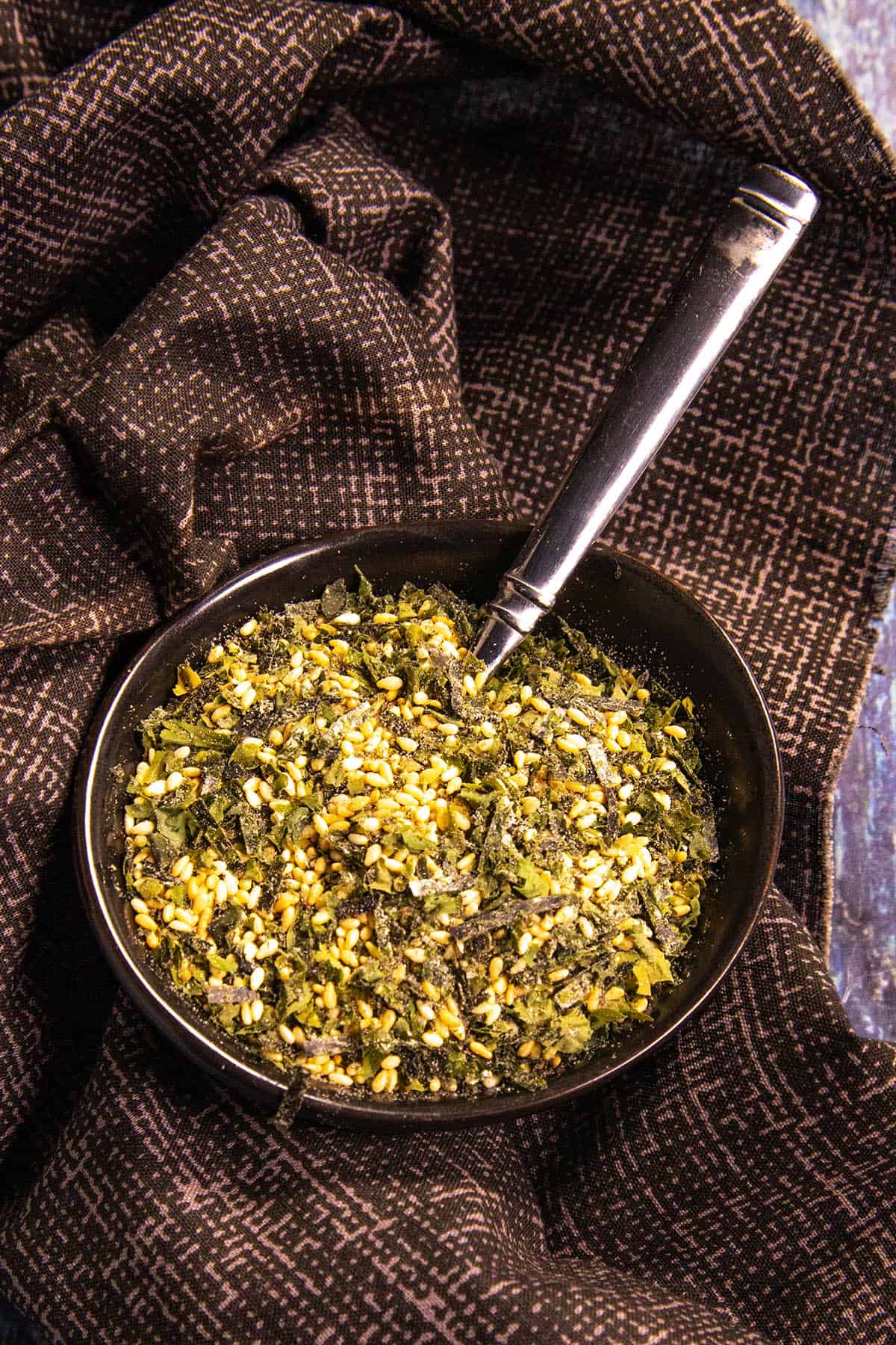 A black bowl filled with vibrant green and yellow Homemade Furikake Seasoning sits on a dark patterned cloth, with a metal spoon resting inside the bowl.