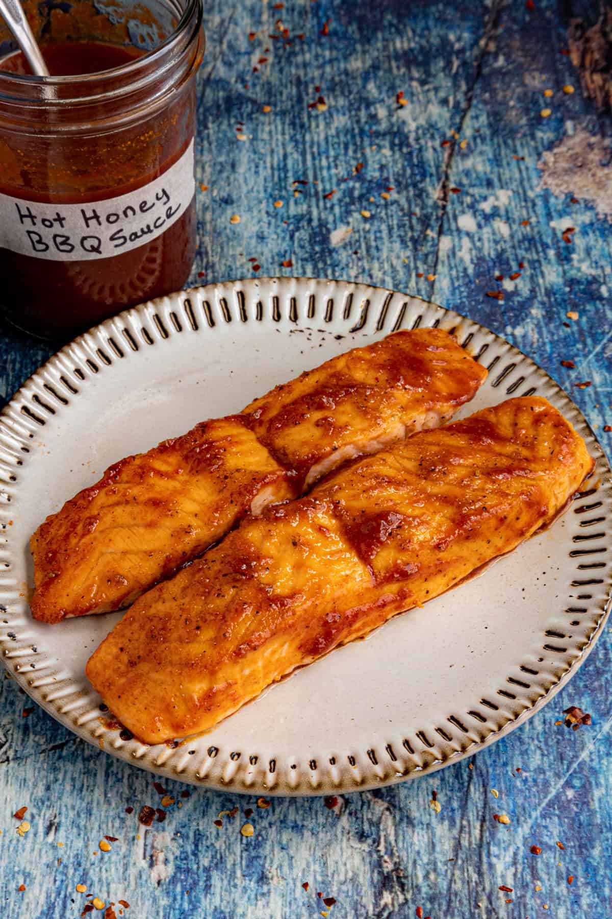 Two cooked salmon fillets coated in a reddish Hot Honey BBQ Sauce sit on a white plate. A jar labeled Hot Honey BBQ Sauce with a spoon inside is in the background on a blue rustic surface.
