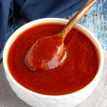 A hand holds a wooden spoon lifting thick, reddish-brown Easy Hot Honey BBQ Sauce from a white bowl, which sits on a cloth napkin atop a blue, textured surface.