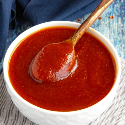 A hand holds a wooden spoon lifting thick, reddish-brown Easy Hot Honey BBQ Sauce from a white bowl, which sits on a cloth napkin atop a blue, textured surface.