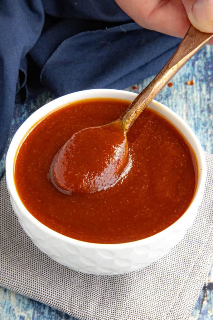 A hand holds a wooden spoon lifting thick, reddish-brown Easy Hot Honey BBQ Sauce from a white bowl, which sits on a cloth napkin atop a blue, textured surface.