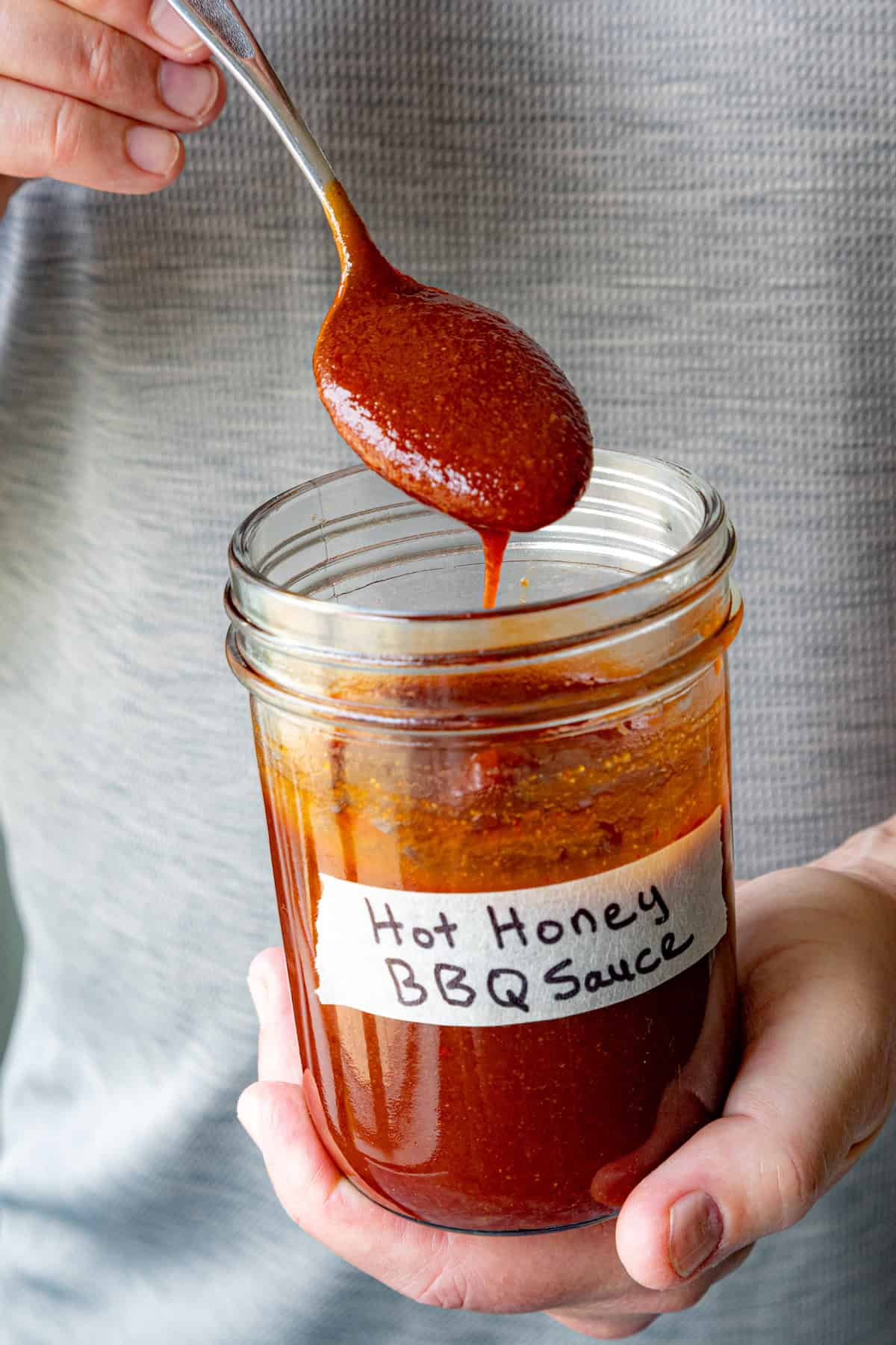 Mike holds a jar labeled Easy Hot Honey BBQ Sauce and uses a spoon to scoop some of the reddish-brown sauce, with sauce dripping back into the jar.