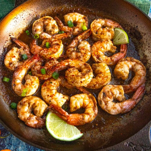 A skillet filled with spicy Jerk Shrimp, garnished with chopped green onions and lime wedges, set on a blue surface with a green cloth nearby.
