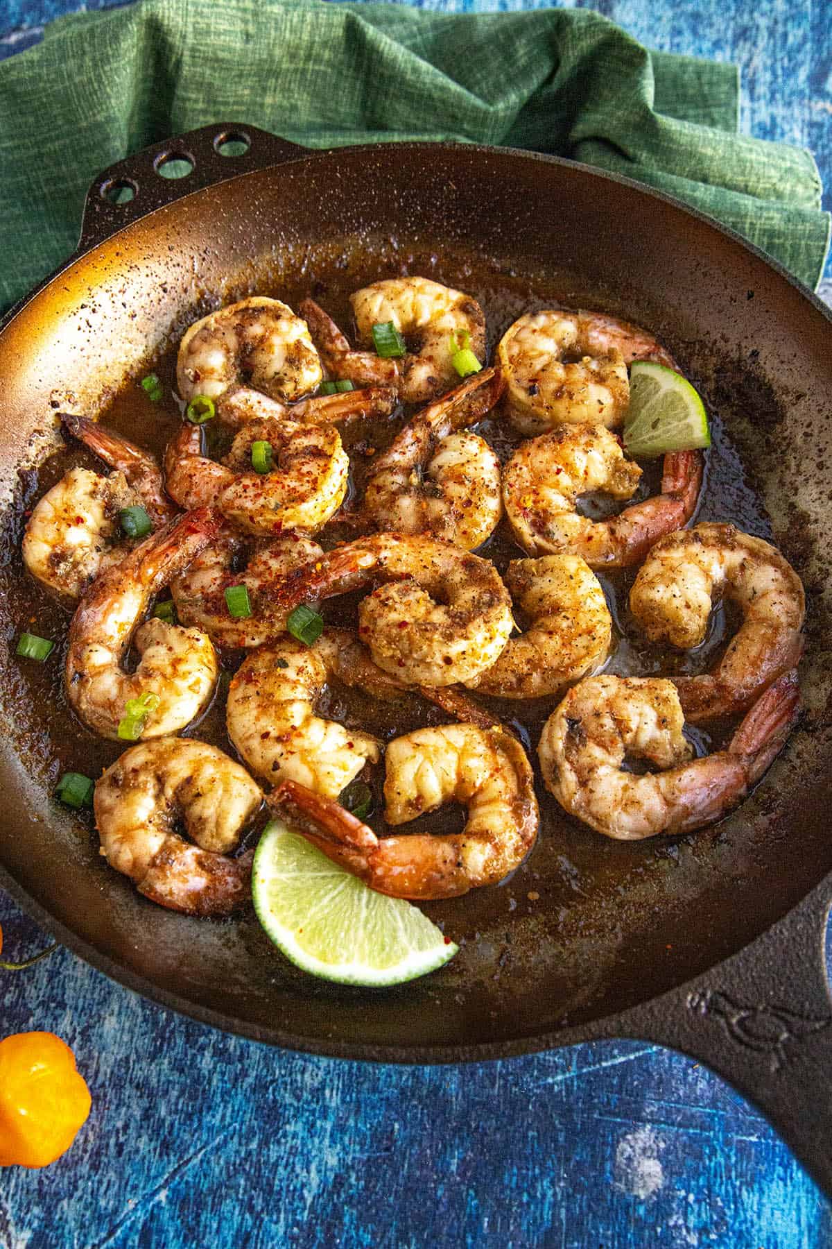A skillet filled with spicy Jerk Shrimp, garnished with chopped green onions and lime wedges, set on a blue surface with a green cloth nearby.